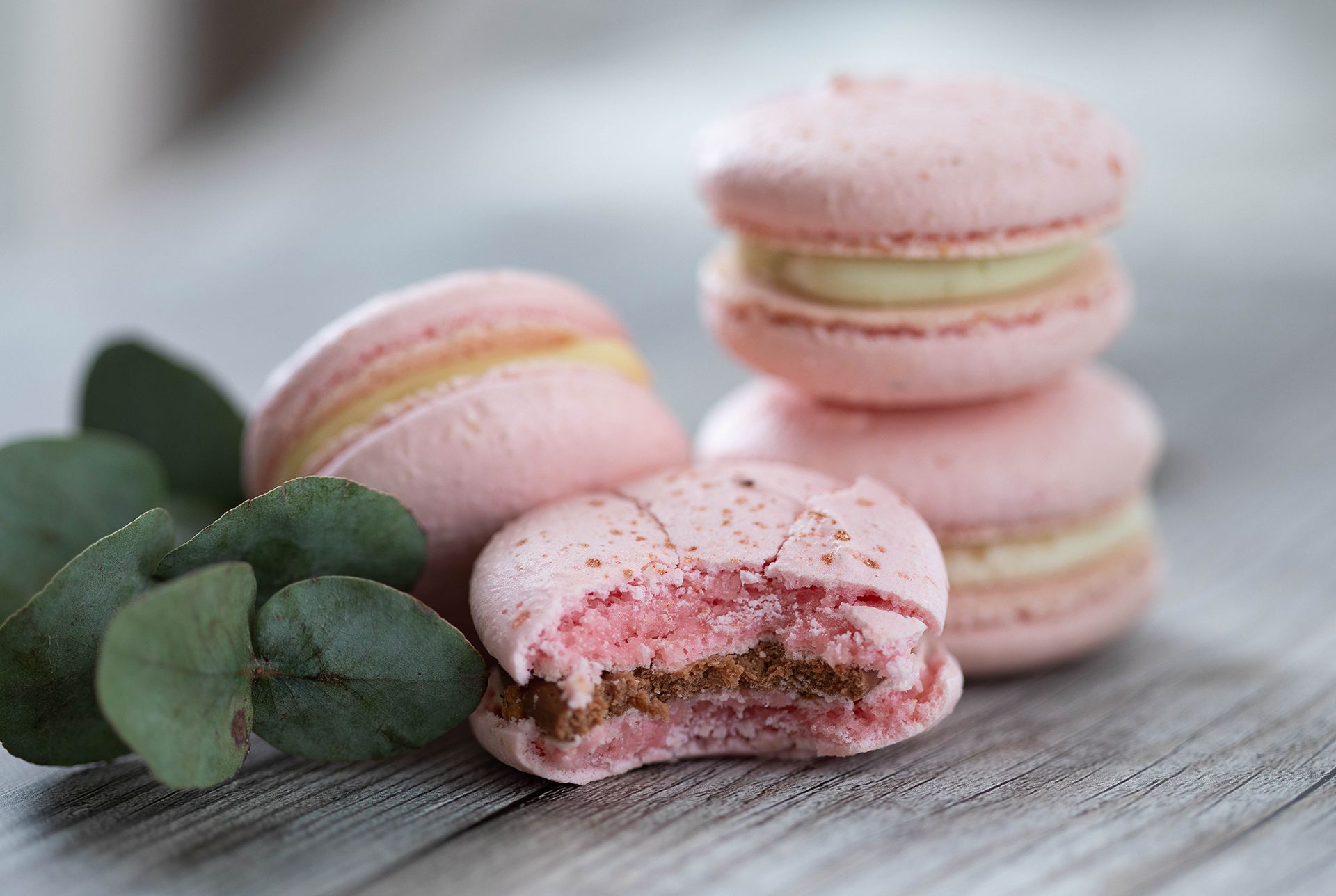 Stack of pink macarons with one half eaten in the fron on a white wooden table next to decorative leaves