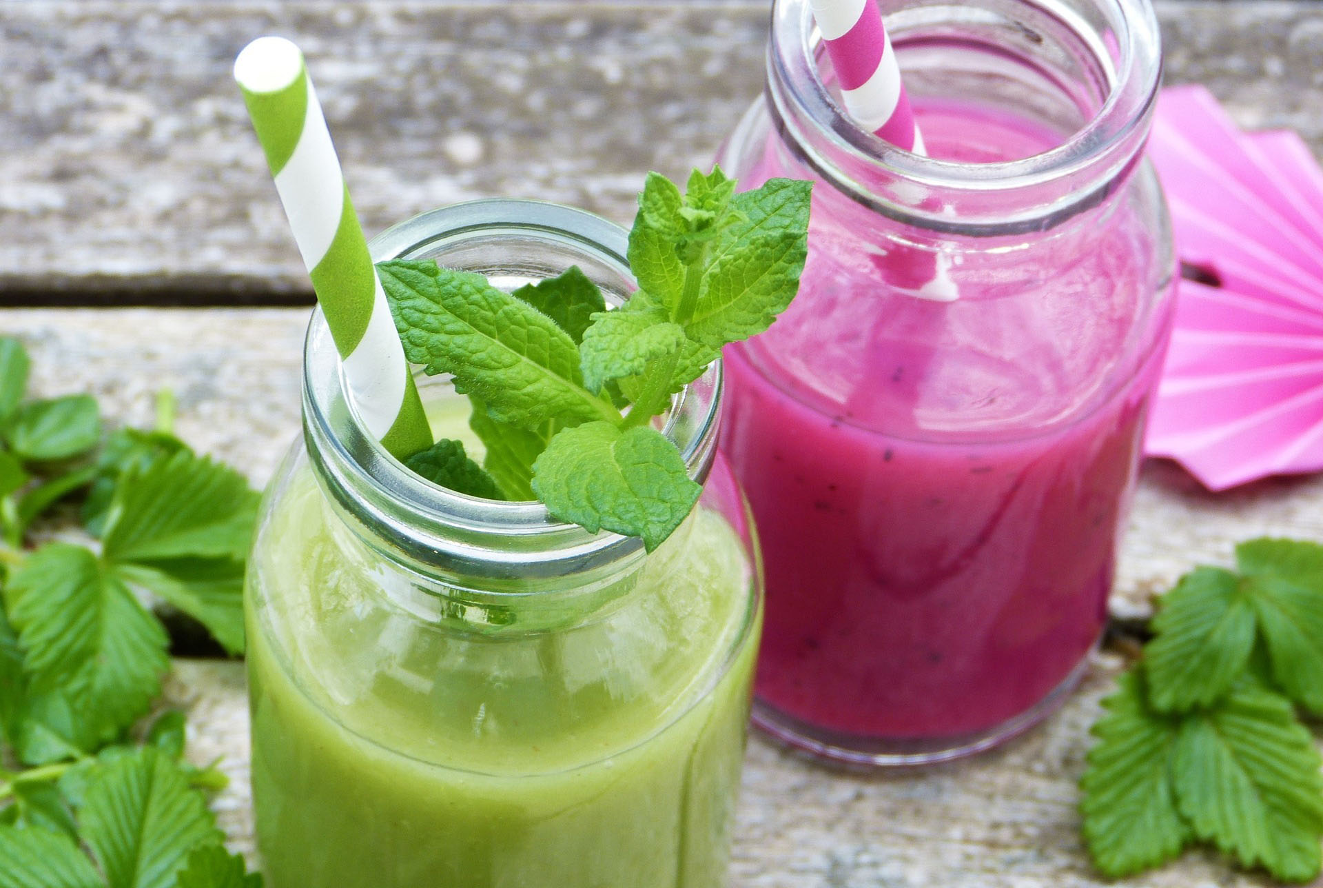 Two glass bottles with  straws filled with smoothies with on wooden table surrounded by mint leaves